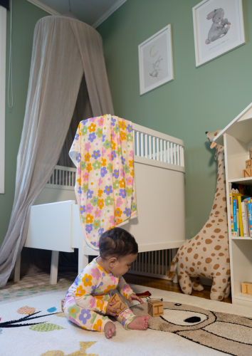 Baby sitting with a bamboo pyjamas playing with toys infront of a crib